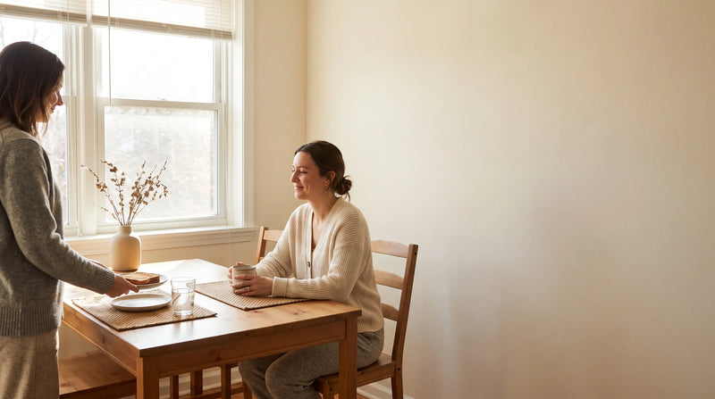 Two friends sharing simple food and water in a warm late-morning apartment kitchen.